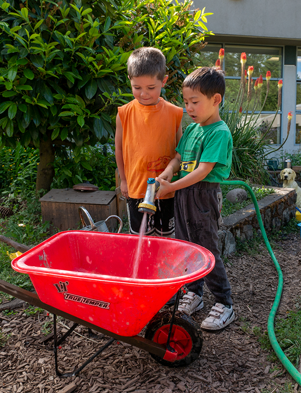 two children filling a wheelbarrow full of water from a hose