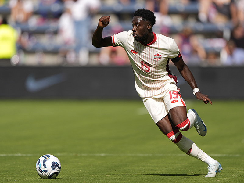 Image 4: Fully body photo of Alphonso Davies kicking a soccer ball in a stadium pitch.
