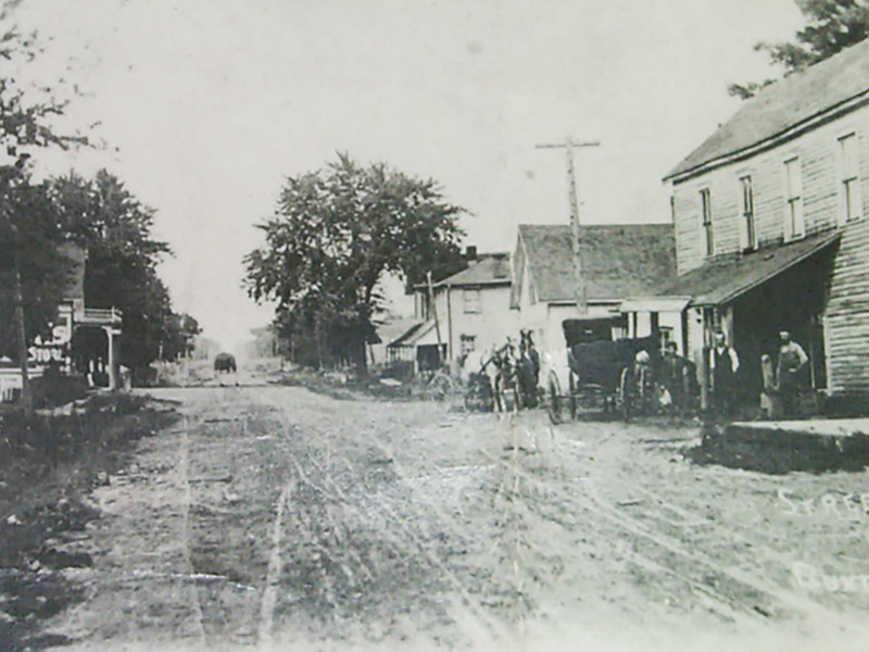 Image 5: Historical black and white photo of wooden buildings with gravel road in front.