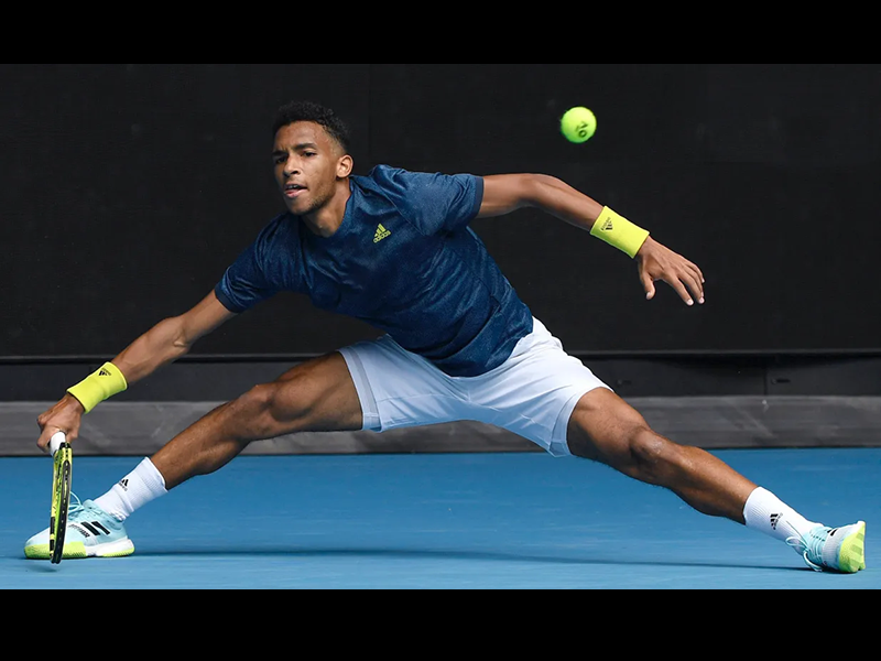 Image 2: Photo of Felix Auger-Aliassime with legs wide apart, holding a tennis racket in one hand and a tennis ball visible in the air in front of him.