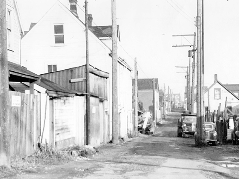 Image 6: Historical black and white photo of wooden buildings beside a road with telephone poles and three old cars parked nearby.