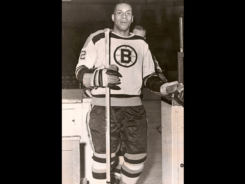 Image 8: Historical black and white full body photo of Willie O’Ree in hockey uniform, holding hockey stick.